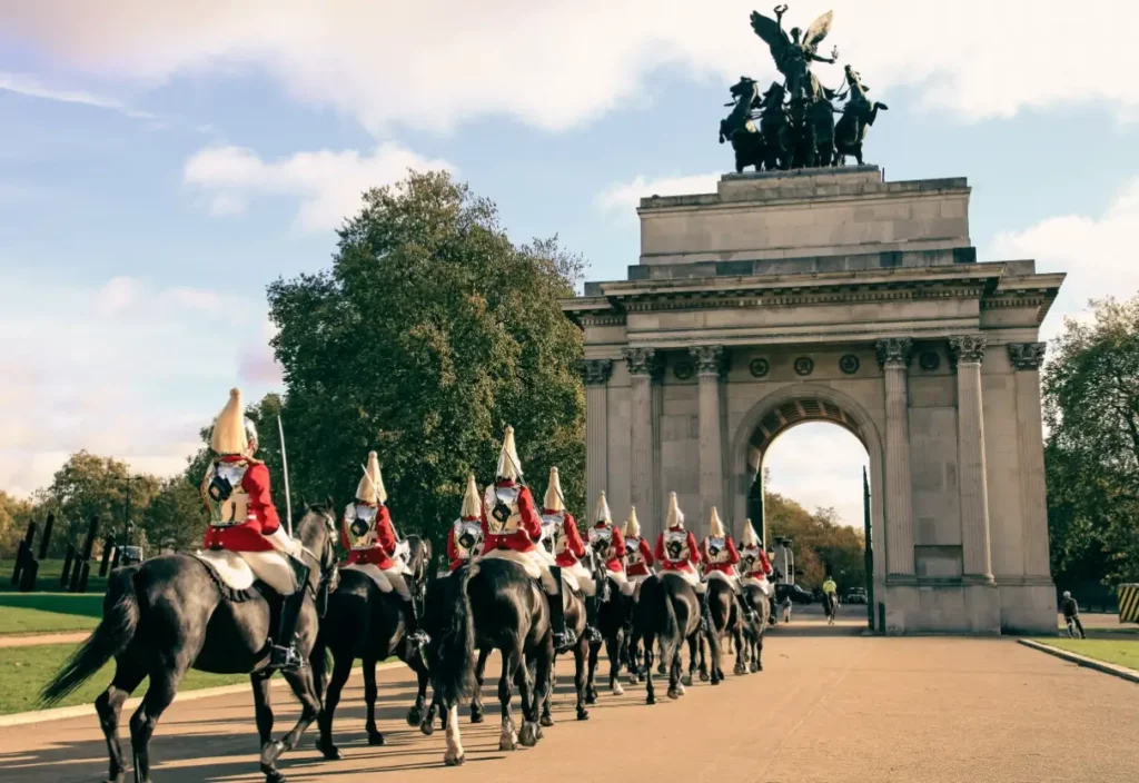 English soldiers on horseback wearing red ceremonial uniforms march through a large stone victory arch in London