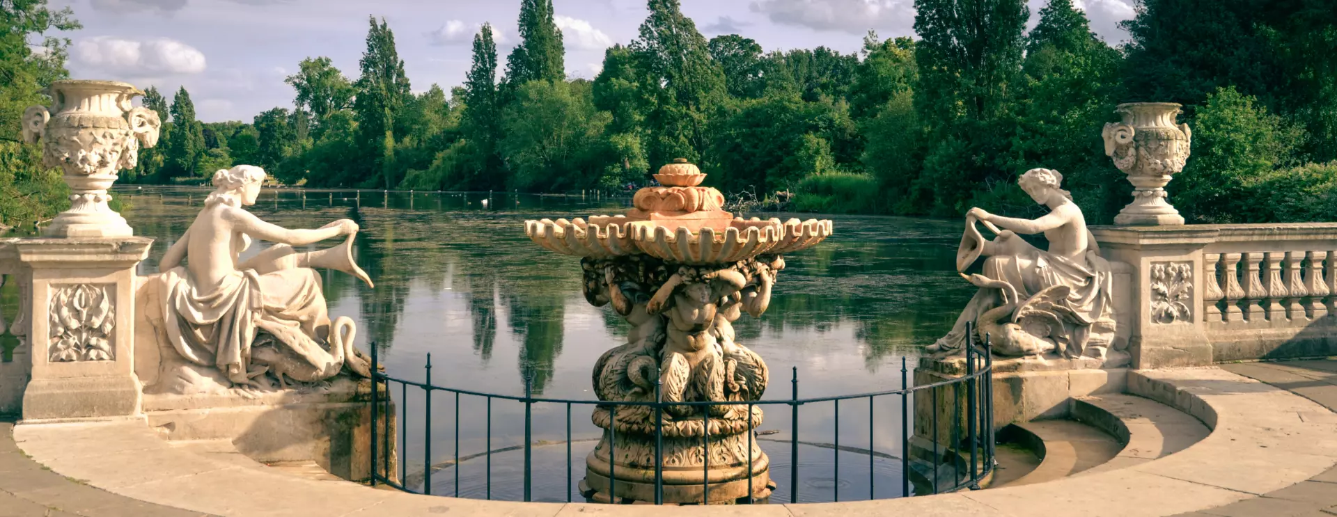 Ornate fountain on a lake surrounded by white stone sculptures and lush green trees