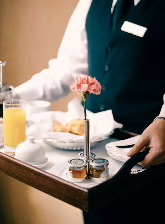 Attendant bringing room service tray with a pastry, jellies, glass of orange juice, and a decorative flower
