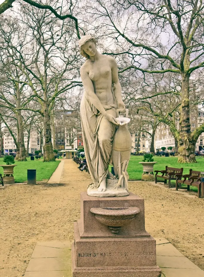 View of a white stone statue of a woman sitting among brown wooden benches in a London city park with green grass
