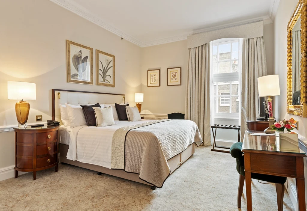 View of stylish bedroom with plush bed with many pillows, side tables with lamps, art on walls, and cream wallpaper